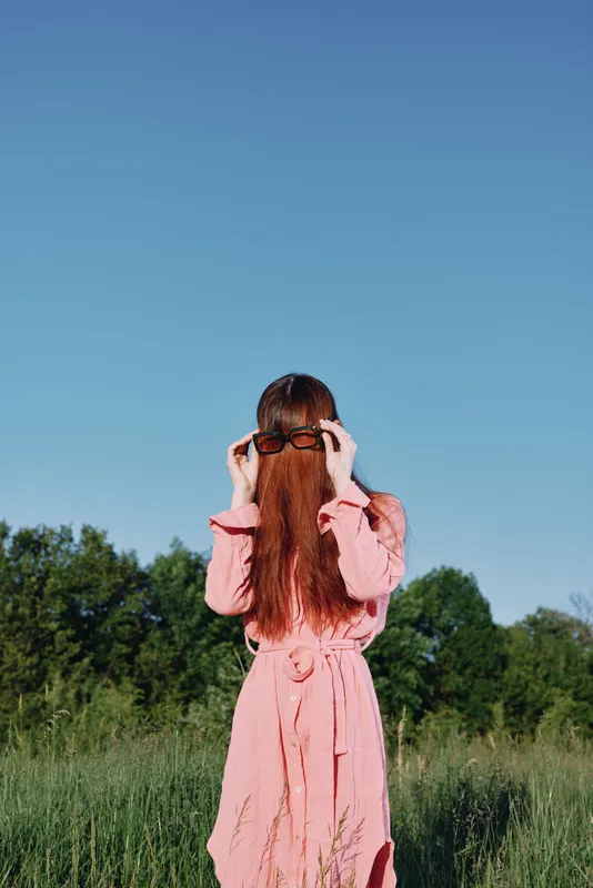 A woman in a pink dress with long hair playfully covering her face, set against a bright blue sky and green field, represents joy and freedom in outdoor fashion