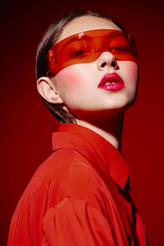 Young woman in red futuristic sunglasses wearing vibrant red outfit looks confidently with bright glossy lipstick and pink blushed cheeks. Bold studio shot for beauty fashion appears modern.