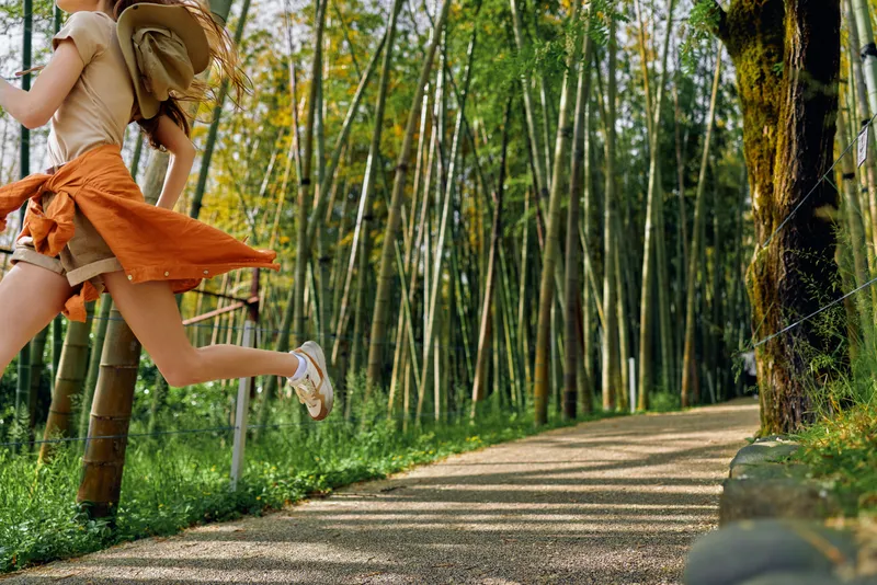 Woman jump in bamboo forest on a winding path wearing a straw hat and orange skirt, summer outdoors scene of walking trail, joyful motion and sunlight filtering through green trees.