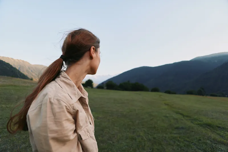 Young woman with long brown hair looks thoughtfully at distant mountains while standing in a wide open grassy field during early evening light.