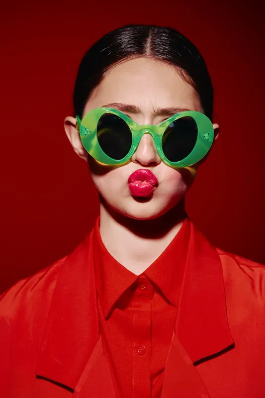 Young woman in bright red shirt wears oversized green sunglasses, puckering vibrant glossy lips. Studio portrait on red background. Concept of beauty, fashion, creative style, bold colors.
