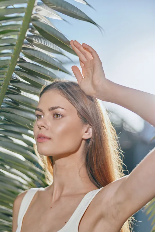 Young woman with long hair poses under palm leaves outdoors, showcasing natural beauty and sunny summer vibes in a bright, clear sky setting.