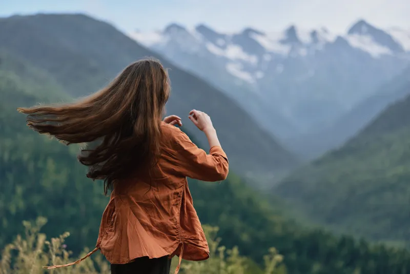 Woman standing in an orange shirt facing majestic mountains, wind in her hair, capturing outdoor adventure and serene nature in a vast valley landscape