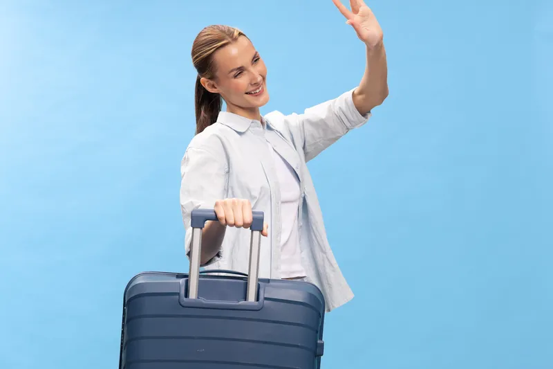Young woman with suitcase waves hand smiling isolated on blue. Concept of travel, vacation, tourism, journey, and friendly greeting with luggage in casual clothing.