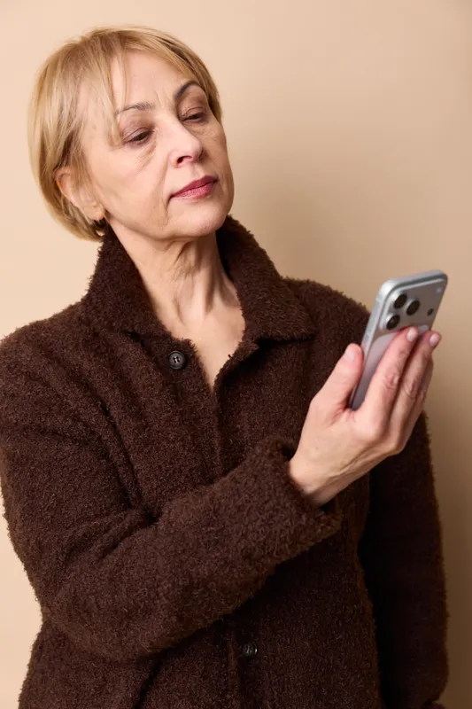 Elderly woman portrait indoors wearing a brown coat holding a smartphone with eyes closed calm expression focused communication by phone on beige background soft warm light