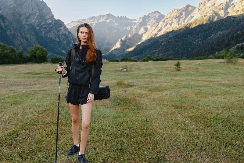 A lone female hiker stands in a wide alpine meadow with a trekking pole, compact bag, and athletic gear, framed by rugged peaks under a clear blue sky.