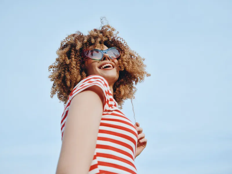 Joyful young woman in a red and white striped shirt smiles under a clear blue sky, photographed from a low angle to emphasize carefree energy and sunny mood.
