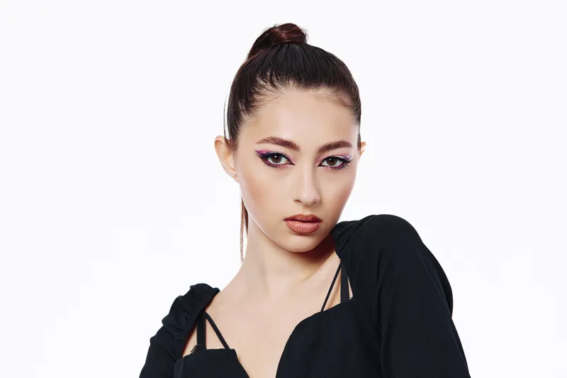 Young woman with creative eye makeup poses looking confidently at camera. Modern black top contrasts pastel purple eyeshadow. Clean white background. Concept of beauty, fashion, and makeup artistry.