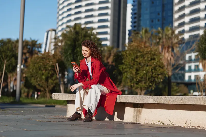 Young woman in a red coat sits on a concrete bench in a sunny urban plaza, smiling while checking her phone, with modern buildings in the background.