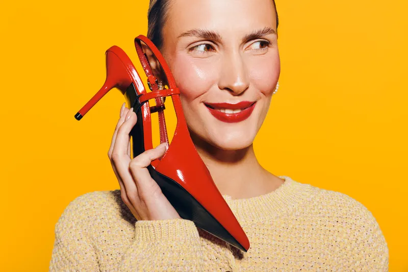 Smiling woman holds red high heel shoe near face on yellow background. Concept of fashion, style, footwear, and beauty with bright colors and cheerful mood in studio lighting.