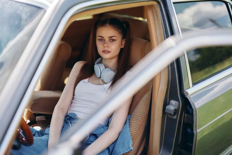 Young woman sitting in a car with headphones, wearing casual clothes, expressing relaxation and joy, surrounded by nature in soft sunlight.