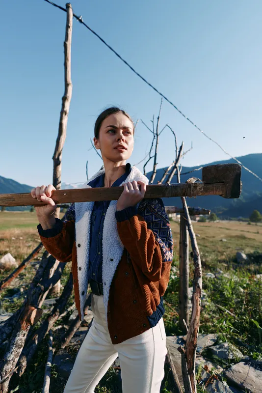 Woman holding axe on shoulder by wooden fence in rural countryside portrait, standing outdoors with mountains in background. Strong pose, rustic landscape and farm atmosphere.