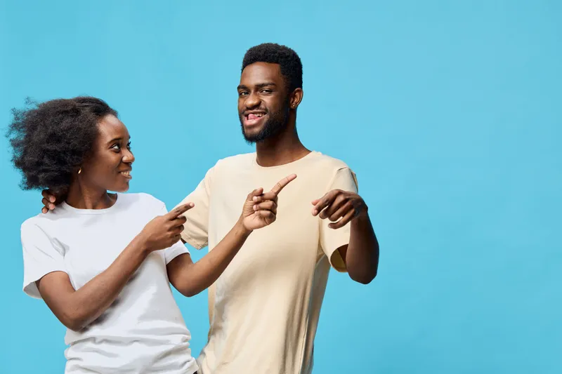 Young couple smiling and pointing at each other against a bright blue background, showcasing joy and connection in a playful moment