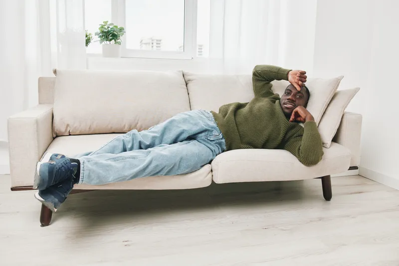 Young Black man relaxing on a modern sofa at home, wearing casual green sweater and jeans, conveying a sense of comfort and leisure in a bright, minimalist living space