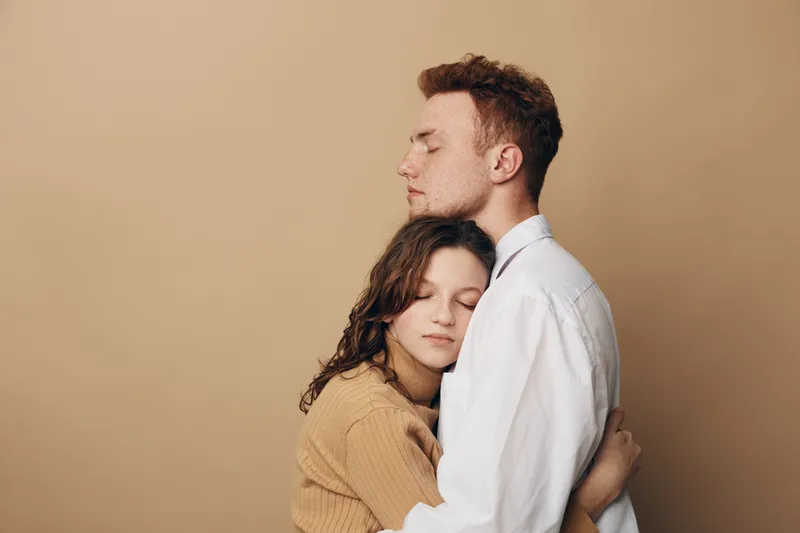 Young couple embraces tenderly with closed eyes, expressing love and comfort. Neutral background highlights emotional connection and warmth between man and woman in casual clothing.