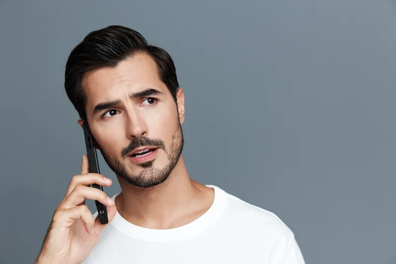 Thoughtful man talking on mobile phone with serious expression, wearing casual white t-shirt, set against plain gray background. Communication concept.