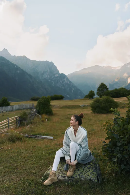 Young woman with brunette hair sits on a rock in a mountain meadow wearing a cozy cardigan and boots, gazing softly into the distance in a tranquil outdoor setting.