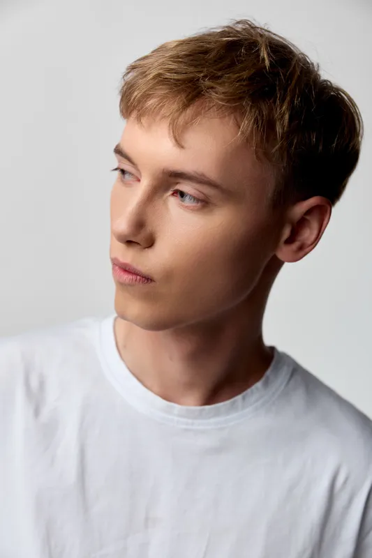 Young man with light hair in a white t shirt gazing to the side in a clean studio setting, thoughtful mood, minimal wardrobe, calm confidence and contemplative attitude.