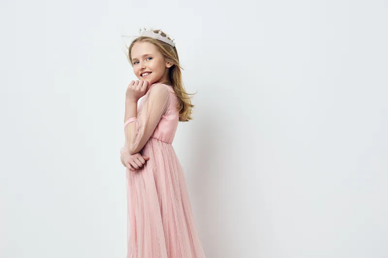 Charming young girl in a pink dress with a crown, smiling joyfully against a minimalistic white background, perfect for seasonal children s fashion themes
