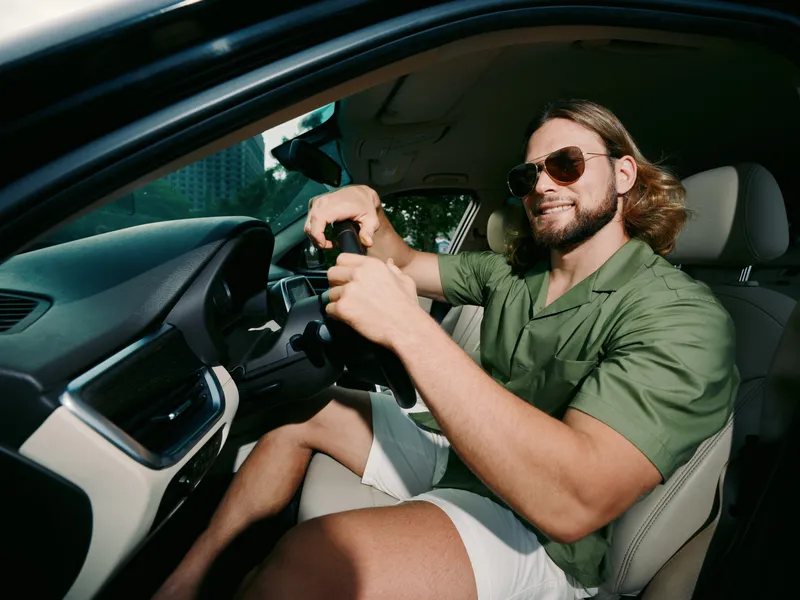 A relaxed man sits in the driver's seat of a modern car, wearing sunglasses and a green short sleeve shirt, smiling confidently while gripping the steering wheel.