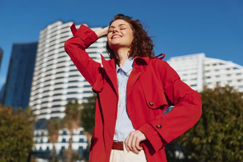 Young woman with curly hair wearing a red coat enjoys sunshine outdoors with eyes closed and a joyful smile on her face.