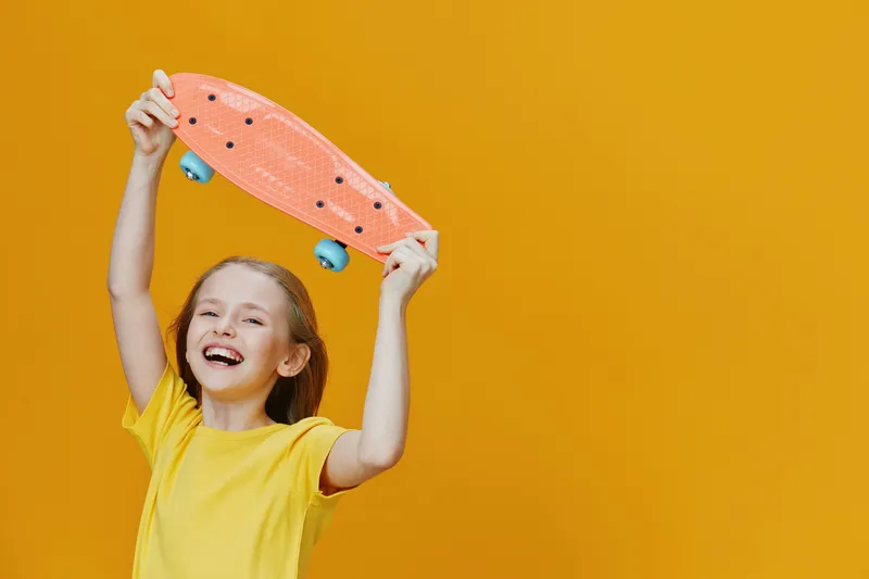 Cheerful girl holding a skateboard against a vibrant yellow background, showcasing joy and youthful energy, reflecting a fun and active lifestyle
