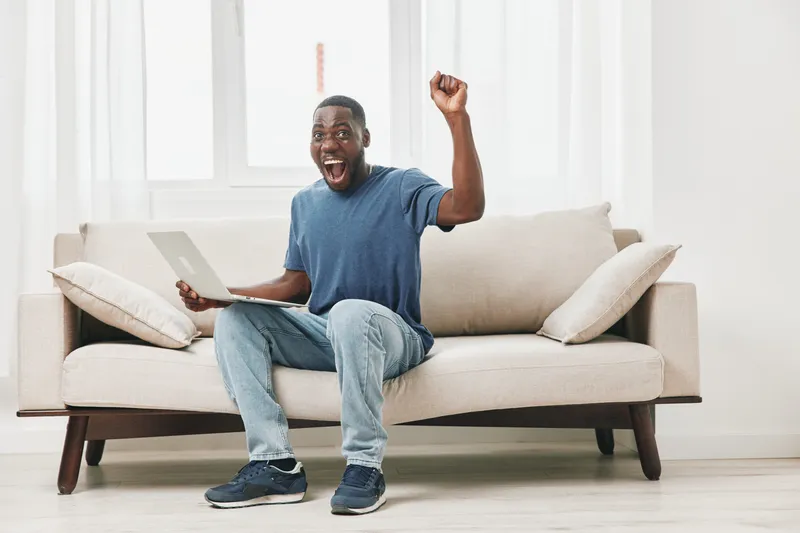 Happy African man celebrating success while using a laptop on a beige sofa, bright room with natural light, casual wear, modern interior