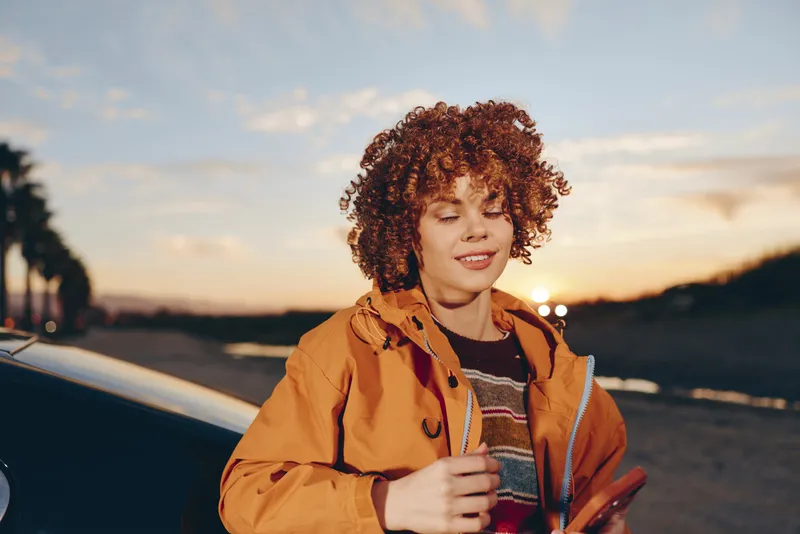 Smiling woman outdoors lifestyle, curly hair in warm sunset light, holding a coffee cup beside a parked car, wearing a brown jacket, relaxed posture, golden evening glow