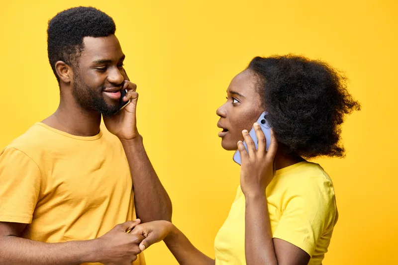 Young African couple engaged in a joyful conversation on their phones, dressed in matching yellow shirts against a vibrant yellow background