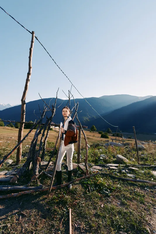 Woman in traditional outfit stands by rustic wooden fence in a rural mountain field, carrying a leather satchel and looking toward distant peaks under clear blue sky.