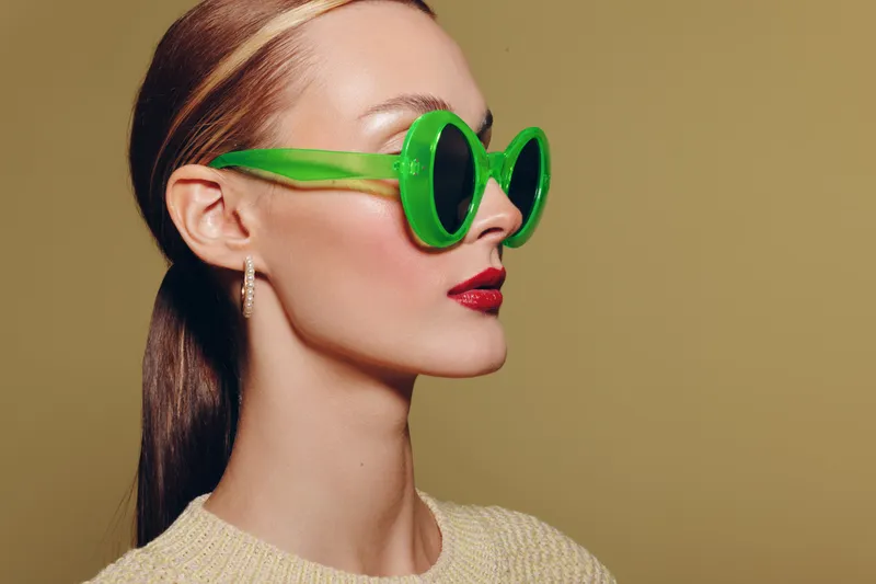 Stylish woman with green sunglasses and red lipstick looks sideways on beige background. Concept of fashion, beauty, accessories, and modern style with minimalistic studio lighting.