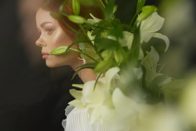 Ethereal portrait of a model with a serene expression surrounded by white lilies against a dark background, highlighting beauty, tranquility, and nature
