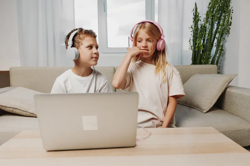 Two children with headphones sit on a couch using a laptop in a bright living room with natural light and green plants in the background.