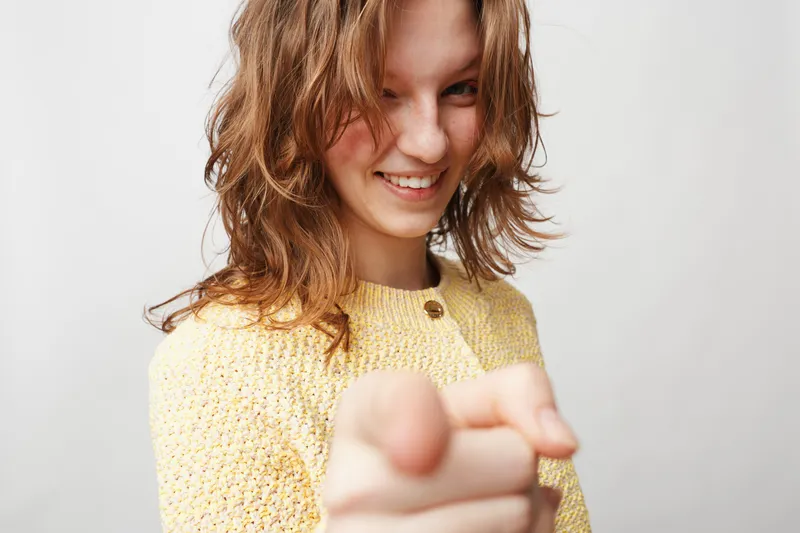 Smiling young woman with wavy hair wearing yellow sweater points finger at camera. Studio portrait on white background. Concept of confidence, positivity, and direct communication.