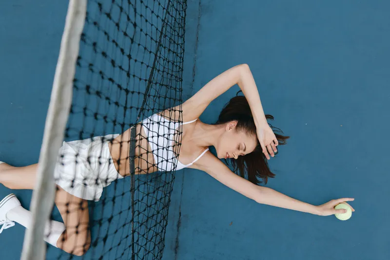 Young female athlete resting on a tennis court, expressing fatigue after a game, wearing a sports bra and shorts, with a vibrant blue background and tennis net