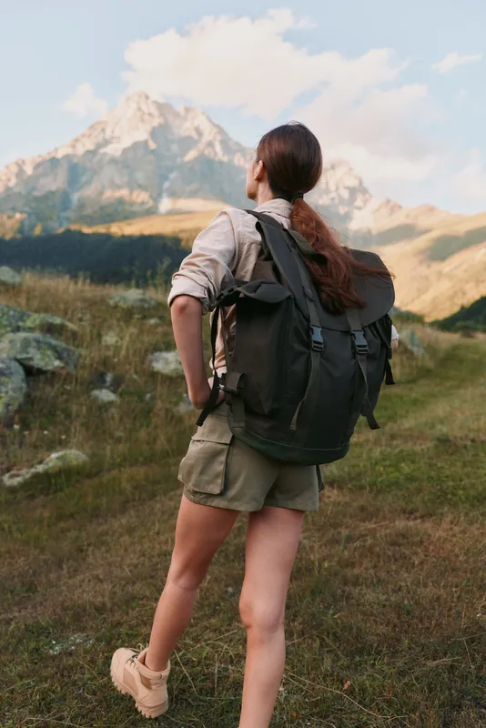 Joyful woman hiking outdoors with backpack, facing distant mountains, sunlit trail, casual attire, sense of exploration, wind in hair, vibrant natural colors, serene wilderness