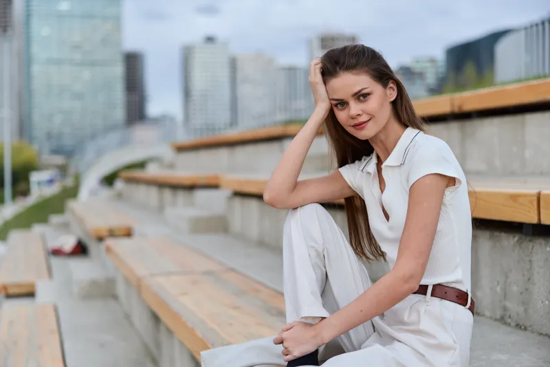 Young woman in white casual outfit sitting thoughtfully on outdoor wooden steps with urban cityscape background, natural light and relaxed mood