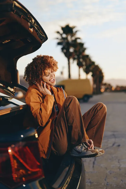 woman lifestyle by car trunk, curly haired woman smiles while talking on phone at sunset, travel mood with casual jacket, warm evening outdoor light, ready to depart