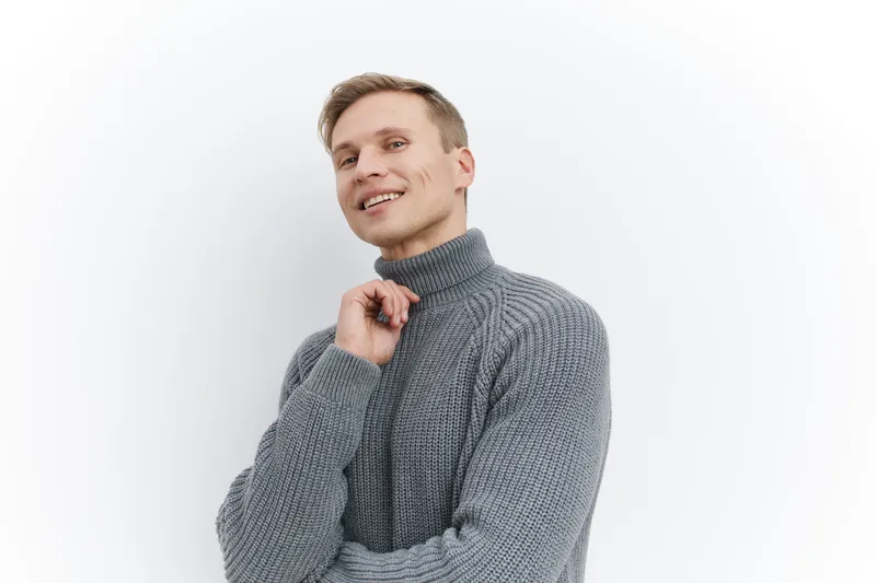 Smiling man in gray turtleneck sweater looking away with hand near chin pose isolated on white background studio portrait. People lifestyle concept