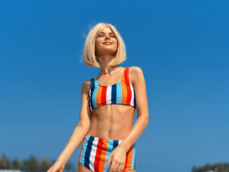 young woman in colorful striped bikini standing outdoors under clear blue sky, smiling and enjoying warm sunny weather, summer beach vibe