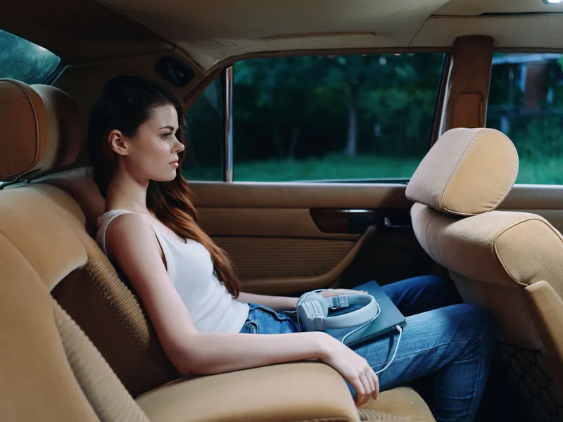 Young woman sitting in a car's back seat, wearing casual attire, reflecting contemplative emotions against a blurred green background.