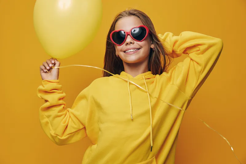 Girl in yellow hoodie holds yellow balloon with smile and heart shaped sunglasses on orange background. Happy child with casual clothes and cheerful expression in studio.