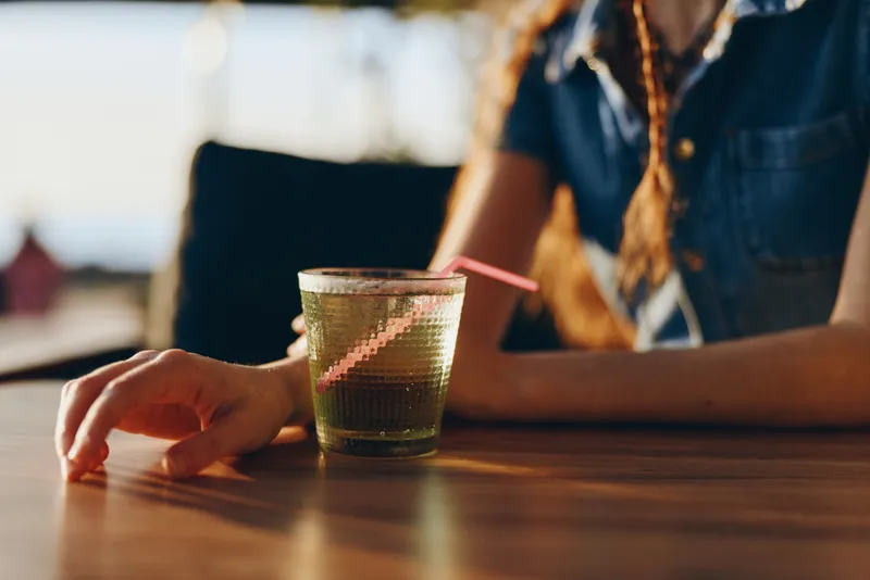 Woman with relaxed hand holding cold refreshing drink with straw on wooden table in bright casual setting, warm light and casual summer atmosphere.
