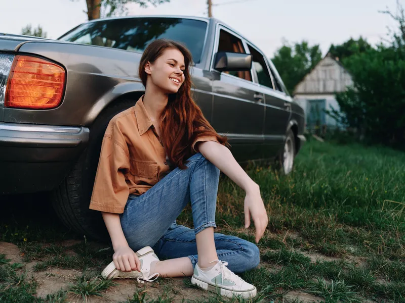 Young woman sitting next to a vintage car, smiling joyfully in casual outfit, showcasing a relaxed summer vibe in nature.