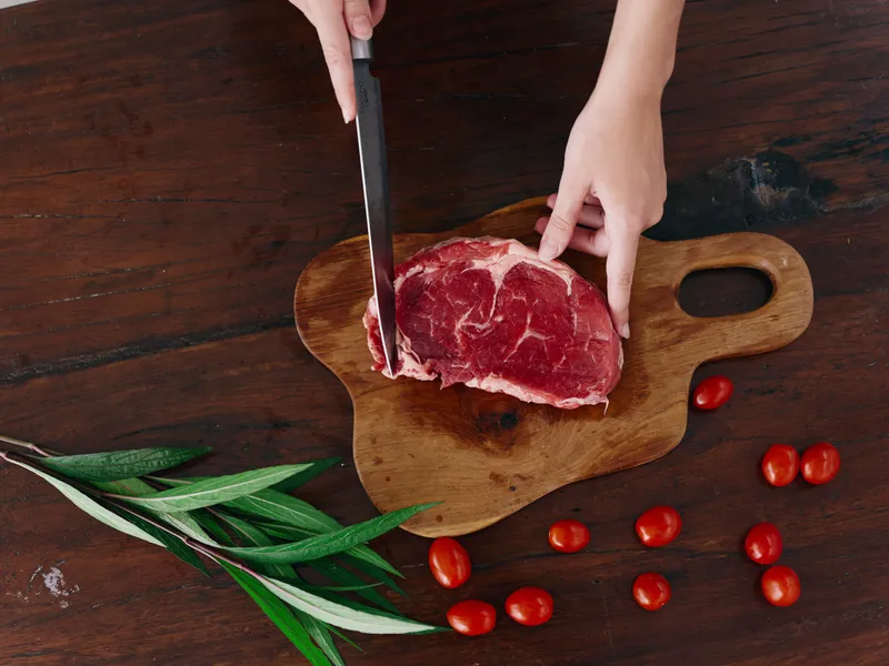 Женщина с ножом в руке нарезает свежее мясо стейка для жарки на кухне