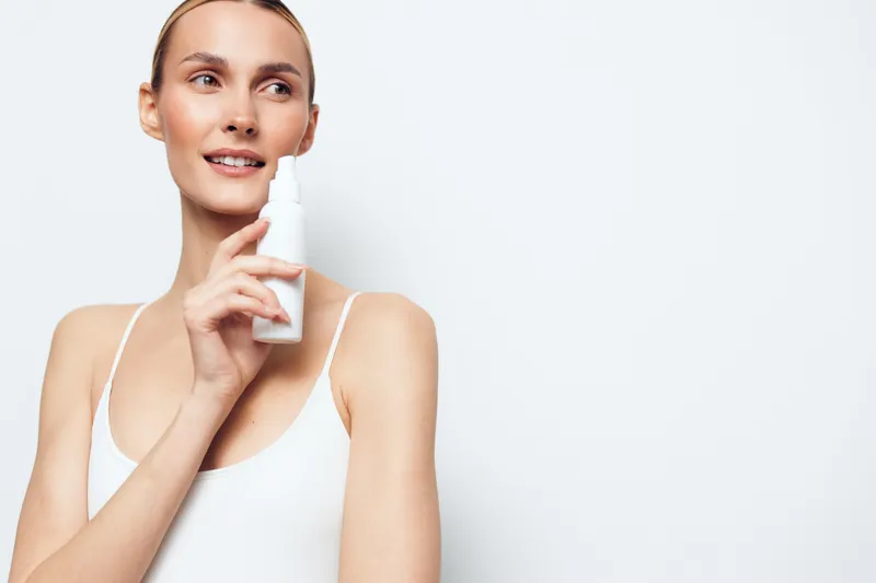 Young woman with clear skin holds nasal spray bottle near face looks away smiling wears white tank top isolated on white background healthcare concept hygiene routine.