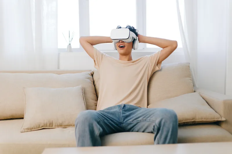 Молодой человек наслаждается виртуальной реальностью дома: он в шлеме VR сидит на уютном диване
