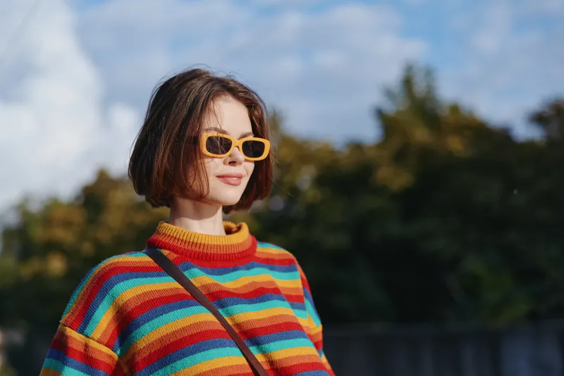 Woman in a colorful striped sweater and yellow sunglasses smiling outdoors, young adult with bob haircut wearing a casual outfit and crossbody bag, lifestyle travel portrait.