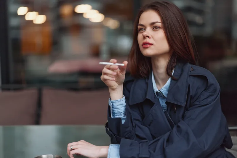 Young brunette woman wearing a blue trench coat and denim shirt sits at a cafe table, smoking a cigarette, and gazing pensively into the distance in a moody urban setting.