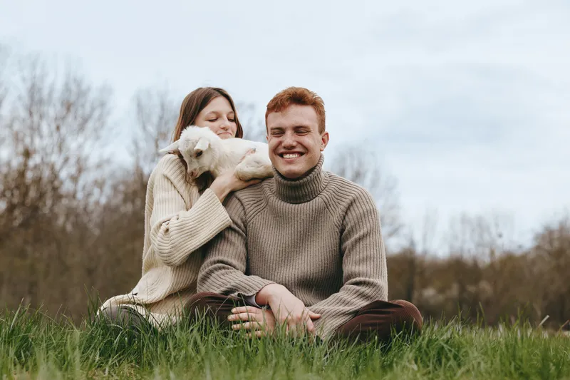 Couple outdoors with a fluffy dog, wearing cozy sweaters, sharing a warm moment in a sunlit field, relaxed posture, gentle breeze, natural colors, lush grass, candid lifestyle, affectionate energy,